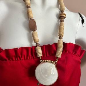 Op de afbeelding: Een lange ketting met een grote, witte schelpenhanger en een goudkleurige rand. De ketting heeft een streng van houten kralen in verschillende vormen en maten, waaronder cilindrische en ovale kralen, en een paar donkerbruine houten kralen. De ketting wordt gepresenteerd tegen een rode achtergrond.