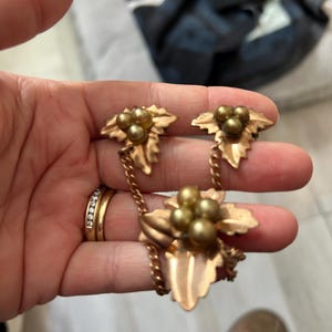 May include: A gold-toned jewellery set featuring a necklace and matching earrings. Each piece has a leaf design with small, round, gold-coloured beads. The necklace and earrings are connected by delicate chains.