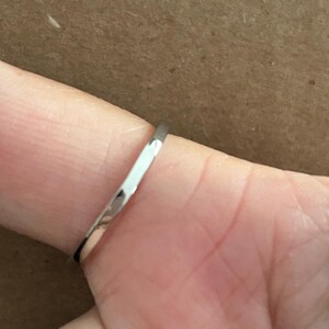 May include: A thin, polished silver-colored metal ring band with a smooth, rounded surface. The band has a reflective finish that catches the light, showing a simple, minimalist design without any stones, engravings, or additional decorative elements.