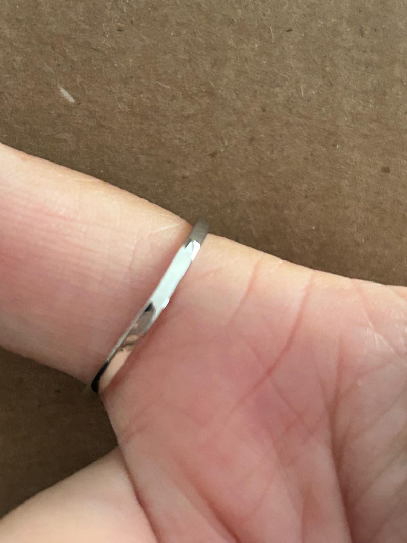 May include: A thin, polished silver-colored metal ring band with a smooth, rounded surface. The band has a reflective finish that catches the light, showing a simple, minimalist design without any stones, engravings, or additional decorative elements.