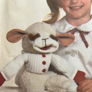May include: A plush lamb doll with a white body, a white sweater with red buttons, and red gloves. The lamb has brown ears and eyes, and a smiling red mouth. The doll is being held by a young girl.