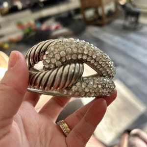 May include: Silver bracelet with a linked design. One section is covered in small, clear crystals, the other has a ribbed texture. The bracelet is held in a hand, highlighting its sparkling details.