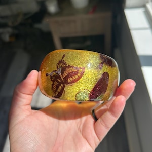 May include: A wide, circular bangle bracelet made of clear resin with a vibrant gold glitter base. The surface is decorated with dark red and brown butterfly motifs and circular patterns embedded within the material, creating a layered effect.