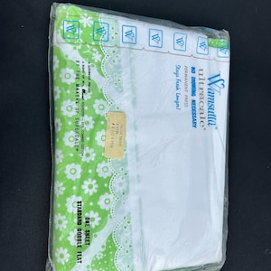 May include: A packaged, flat double sheet from Wamsutta Ultracale. The sheet is white with a green floral patterned border and white lace trim. The packaging includes text that reads "No Ironing Necessary" and "Stays Fresh Longer!"