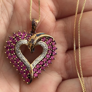 May include: A gold-toned necklace featuring a heart-shaped pendant. The pendant has a silver-colored heart outline, surrounded by small, pink gemstones. The necklace chain is a delicate gold color.