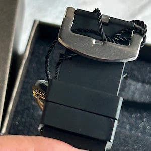 May include: A black silicone watch strap featuring a metallic buckle and a black string tag attached to the hardware. The strap has two sliding loops and is positioned within a dark, textured box, showing a glimpse of a gold-toned circular element.