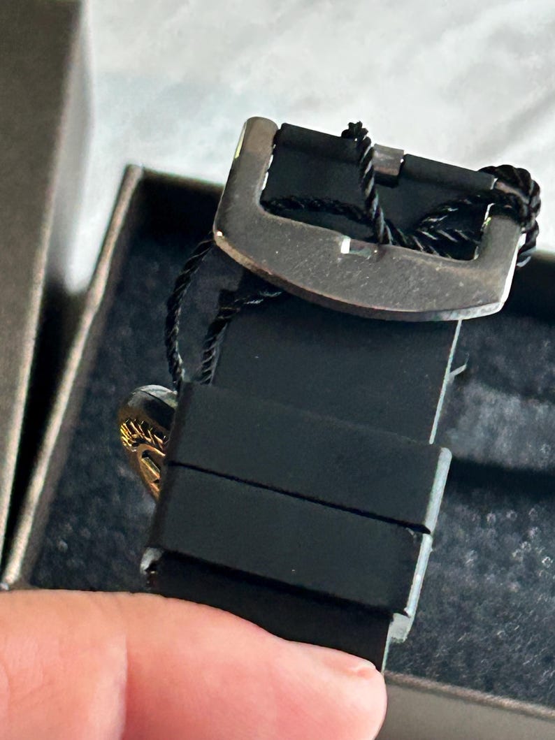 May include: A black silicone watch strap featuring a metallic buckle and a black string tag attached to the hardware. The strap has two sliding loops and is positioned within a dark, textured box, showing a glimpse of a gold-toned circular element.