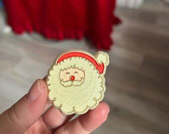 Può includere: Una spilla a forma di Babbo Natale con viso e barba color crema, cappello rosso e naso rosato. La spilla ha un bordo smerlato ed è tenuta in mano.