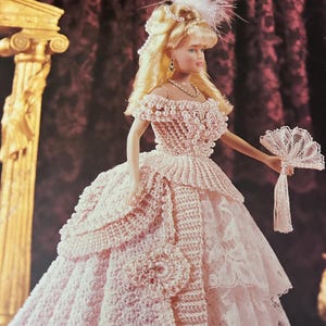 May include: A doll in a pink crocheted gown with a layered skirt and a matching fan. The doll has blonde hair and is wearing a pearl necklace and earrings. The image is from a book titled "Crochet Collector Costume Volume 42."