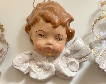 May include: A vintage angel ornament with a cherubic face, rosy cheeks, and light brown hair. The angel's wings are white with iridescent glitter. The ornament is likely made of ceramic or a similar material, and is designed for hanging.
