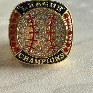 Peut inclure: Une bague de champion dorée avec un motif de baseball central incrusté de strass transparents et de coutures rouges. La bordure noire affiche le texte LEAGUE en haut et CHAMPIONS en bas, avec des pierres rouges sur les côtés.