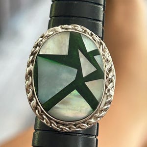 Puede incluir: Un anillo ovalado con un borde trenzado de plata y una incrustación central de piezas de nácar blanco iridiscente separadas por líneas geométricas de color verde oscuro. El anillo está colocado sobre un medidor de anillos de plástico negro.