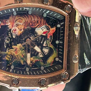 May include: A rose gold-toned wristwatch with a tonneau-shaped case featuring a black dial. The dial displays a detailed graphic of a tiger and a dragon, with visible mechanical gears, white numerical markers, and a black silicone strap.