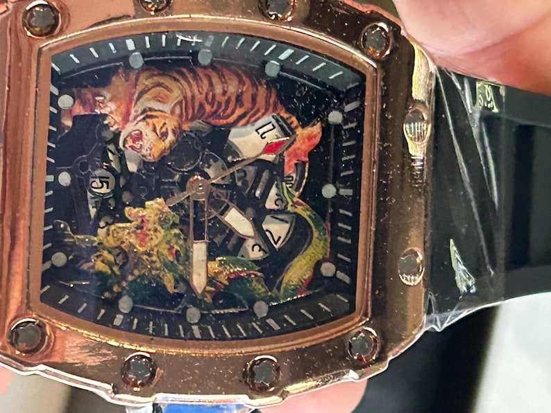 May include: A rose gold-toned wristwatch with a tonneau-shaped case featuring a black dial. The dial displays a detailed graphic of a tiger and a dragon, with visible mechanical gears, white numerical markers, and a black silicone strap.