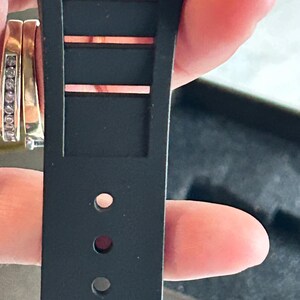 May include: A black watch strap made of a flexible material featuring a series of rectangular cutouts along the upper portion and a vertical row of circular adjustment holes along the lower section, designed for secure fastening to a timepiece.
