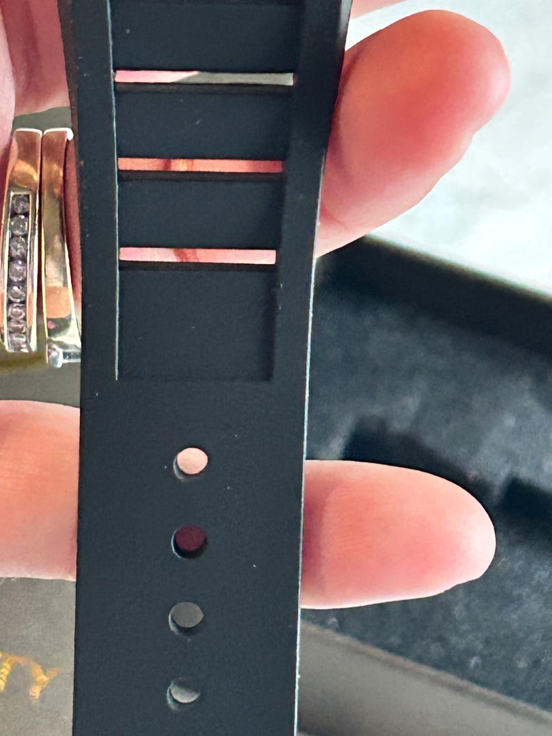 May include: A black watch strap made of a flexible material featuring a series of rectangular cutouts along the upper portion and a vertical row of circular adjustment holes along the lower section, designed for secure fastening to a timepiece.