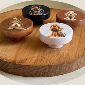 May include: Four decorative wooden knobs with animal illustrations. The knobs are in various colours: dark brown, black, white, and light brown. Each knob features a different dog or cat design. The knobs are displayed on a round wooden surface.