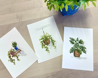 May include: Three watercolour paintings of potted plants. The first painting shows a hanging plant with green leaves and a brown pot. The second painting shows a potted plant with green leaves and a blue pot on top of a stack of books. The third painting shows a potted plant with green leaves and a brown pot.