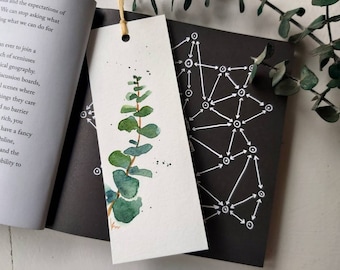 May include: A watercolor painting of a eucalyptus branch with green leaves on a white bookmark. The bookmark is laying on top of a black book with a white geometric pattern.