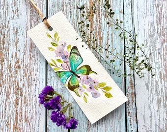 Pode incluir: Um marcador de página de aquarela com uma borboleta verde com marcas marrons, cercada por flores roxas e brancas e folhas verdes.