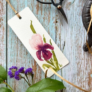 May include: A watercolour bookmark featuring a pink and purple iris flower with green stems. The bookmark is on a light blue wooden surface.