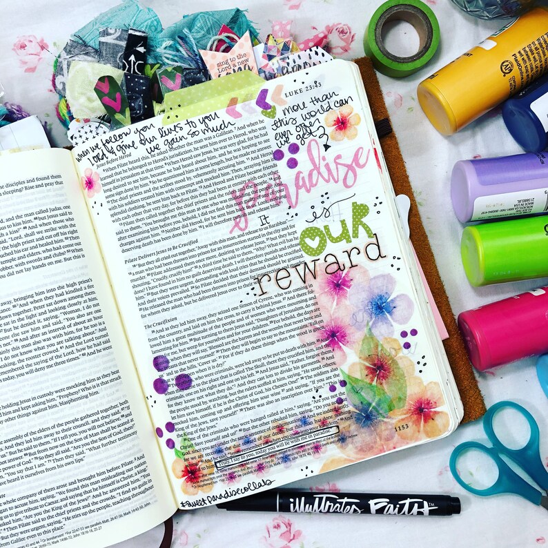 Puede incluir: Biblia abierta con ilustraciones florales coloridas y texto manuscrito, incluyendo las palabras "Paradise is our reward". Tambi&eacute;n se ven suministros adicionales como marcadores, cinta washi y tijeras.