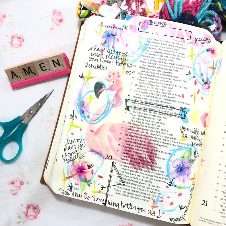 Puede incluir: Un libro abierto con ilustraciones florales coloridas y notas manuscritas. Un flamenco rosa est&aacute; dibujado en la p&aacute;gina, con el texto "Know there is something better for us!" y "Amen" en un bloque de madera.