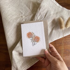 Op de afbeelding: Een wenskaart met een aquarel illustratie van twee oranje en gele protea bloemen. De kaart wordt tegen een neutrale linnen doek en een bruine kartonnen achterkant gehouden. Gedroogd konijnenstaartgras staat op de achtergrond.