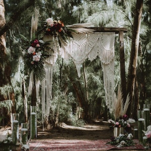 May include: A rustic wooden archway decorated with a white macrame tapestry and floral arrangements. The archway is set in a lush green forest setting.
