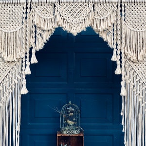 May include: A white macrame archway with a geometric pattern hangs in front of a blue door. The archway is made of thick cotton rope and has a fringe detail.