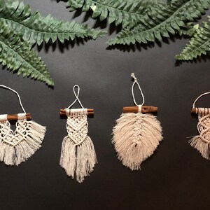 May include: Four handmade macrame wall hangings with wooden dowels. Each hanging features a different design, including a heart, a feather, and two geometric patterns. The hangings are made with natural white cord and have a rustic, bohemian aesthetic.