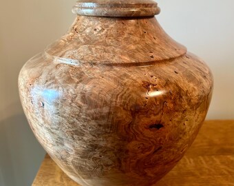 May include: A polished wooden urn with a rounded body and a matching lid. The wood features a complex, swirling burl grain pattern in shades of light brown, tan, and dark amber, showcasing a smooth, glossy finish across the entire surface.