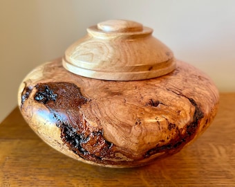 May include: A handcrafted wooden vessel with a rounded, bulbous body featuring prominent natural wood grain patterns, dark knots, and a smooth, polished finish. The container is topped with a matching circular, tiered wooden lid.