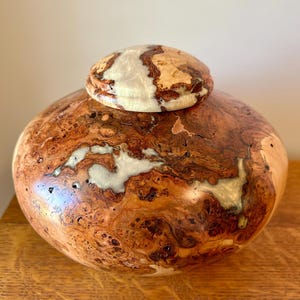 May include: A rounded wooden vessel with a matching lid, featuring a polished finish that highlights the natural brown burl wood grain. The surface is accented with irregular, smooth, pearlescent white resin inlays integrated into the wood.