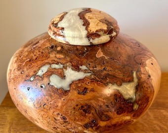 May include: A rounded wooden vessel with a matching lid, featuring a polished finish that highlights the natural brown burl wood grain. The surface is accented with irregular, smooth, pearlescent white resin inlays integrated into the wood.