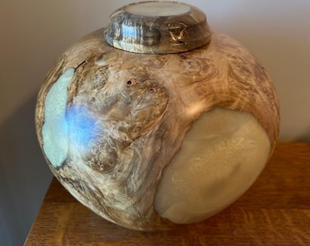 May include: A spherical wooden vessel featuring a polished burl wood grain with natural knots and swirls. The container includes a matching circular lid and is accented with smooth, opaque, light-colored resin inlays.