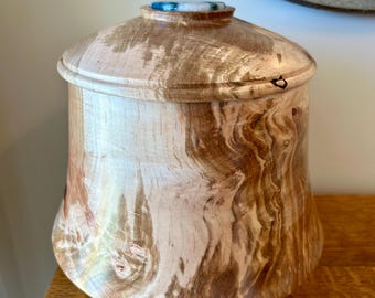 May include: A turned wooden vessel with a smooth, polished finish featuring natural light and dark brown wood grain patterns. The container has a matching lid with a small, circular, blue and white marbled inlay centered on the top surface.