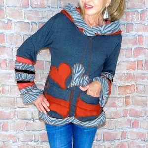 May include: A dark gray hooded jacket with red heart and zebra print accents. The jacket has red pockets and cuffs with black stripes. The person is wearing blue jeans and black sneakers.