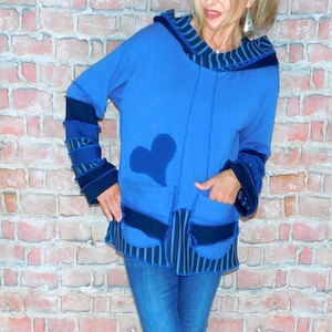 May include: A blue hooded tunic with a heart applique and pocket details. The tunic has dark blue accents on the cuffs, pockets, and hood. The model is wearing blue jeans and black and white sneakers.