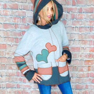 May include: A gray hooded sweatshirt with a heart design and two front pockets. The hood and sleeves have striped accents in shades of brown, teal, and gray. The model is wearing blue jeans.
