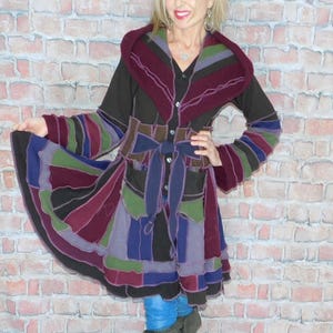 May include: A patchwork coat in shades of black, purple, green, and blue. The coat features a hood, a tie belt, and long sleeves with striped cuffs. The coat is knee-length and has a flared skirt.