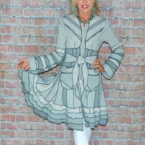 May include: A light gray, long-sleeved, hooded coat featuring a patchwork design with visible seams, a tie-front waist closure, and a flared, ruffled hemline. The coat is paired with white cropped trousers and white slip-on shoes.