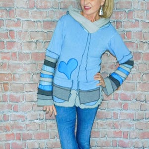 May include: A light blue hooded jacket with a large blue heart applique on the front. The jacket has gray accents on the sleeves and pockets. The person is wearing blue jeans and dark blue sneakers.