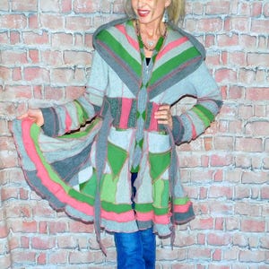 May include: A long, hooded coat with a patchwork design in shades of gray, green, and pink. The coat features a zip-up front, pockets, and a tie belt. The sleeves and hem have a ruffled edge. The person is wearing blue jeans and black sneakers.