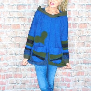 May include: A blue hooded jacket with olive green and black accents. The jacket features large pockets and a unique heart-shaped design. The model is wearing blue jeans and camouflage print shoes. The background is a brick wall.
