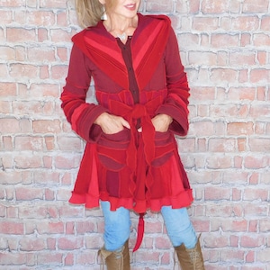 May include: A red hooded coat with a tie waist and pockets. The coat features a layered design with varying shades of red. The person is wearing blue jeans and brown knee-high boots. The background is a brick wall.