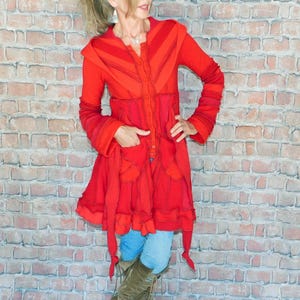 May include: A vibrant red coat with a unique design, featuring a button-up front, pockets, and a flared skirt. The coat has long sleeves and is paired with blue jeans and brown boots. The background is a brick wall.