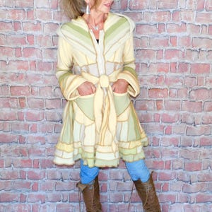 May include: A long, patchwork coat in shades of cream, green, and gold. The coat features a hood, pockets, and a tie belt. The model is wearing blue jeans and brown boots. The coat's design is a mix of geometric shapes and textures.