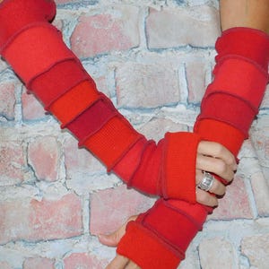 May include: A pair of long, fingerless arm warmers in various shades of red and orange. The arm warmers feature a patchwork construction with visible, raised seams between the horizontal fabric segments, extending from the wrist to the upper arm.