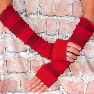 May include: Red arm warmers with a layered design. The warmers are a gradient of red hues, with ribbed cuffs and sections. The background is a brick wall.